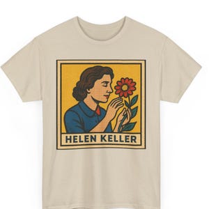 Peut inclure: T-shirt beige avec un graphisme de Helen Keller sentant une fleur rouge. L'illustration est dans un carré jaune avec un bord noir, et le nom "HELEN KELLER" est imprimé en dessous en blanc.