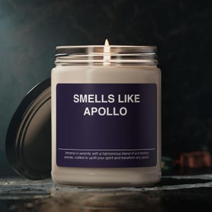 Op de afbeelding: Een glazen kaars pot met een witte kaars erin. De pot heeft een zwart label met witte tekst die "SMELLS LIKE APOLLO" luidt. De tekst onder het label luidt "Immerse in serenity with a harmonious blend of enchanting aromas, crafted to uplift your spirit and transform any space."