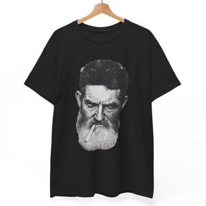May include: Black t-shirt featuring a black and white portrait of a bearded man with a cigarette. The man has a stern expression and a full beard. The t-shirt is hanging on a wooden hanger.
