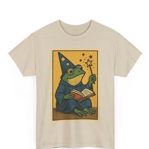 Könnte beinhalten: Beiges T-Shirt mit einer skurrilen Illustration eines grünen Frosches als Zauberer. Der Frosch trägt eine blaue Robe und einen sternenbesetzten Hut, hält einen Zauberstab und ein offenes Buch. Das Design befindet sich in einem gelben Rechteck.