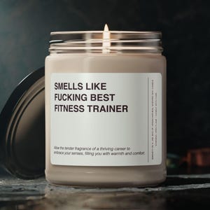 May include: A beige scented candle in a glass jar with a black lid. The label reads "SMELLS LIKE FUCKING BEST FITNESS TRAINER". The candle has a lit wick and is designed to fill a room with fragrance.