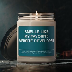 Op de afbeelding: Een witte kaars in een glazen pot met een teal label waarop staat "Smells Like My Favorite Website Developer". Het label bevat ook de tekst "Immerse in serenity with a harmonious blend of enchanting aromas, crafted to uplift your spirit and transform any space."