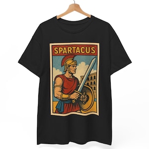 May include: Black t-shirt featuring a vintage-style graphic of Spartacus, a Roman gladiator, holding a sword and shield. The design includes the word "SPARTACUS" in a rectangular frame. The gladiator is depicted in red and gold attire.