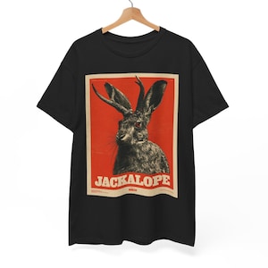 Op de afbeelding: Een zwart t-shirt met korte mouwen en een rechthoekige grafische print op de borst. De print toont een gedetailleerde illustratie van een jackalope tegen een effen rode achtergrond, met het woord JACKALOPE in dikke witte letters.