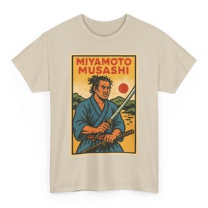 Puede incluir: Camiseta beige con un gráfico de Miyamoto Musashi, un espadachín japonés. La ilustración muestra a Musashi con un kimono azul sosteniendo dos espadas, con un sol rojo y un paisaje de fondo. El texto "MIYAMOTO MUSASHI" está en la parte superior.