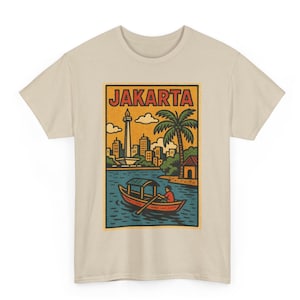 May include: Beige t-shirt featuring a vintage-style illustration of Jakarta. The design includes a boat on water, palm trees, buildings, and the text "JAKARTA" in a rectangular frame. The shirt is made of a soft, light-colored fabric.