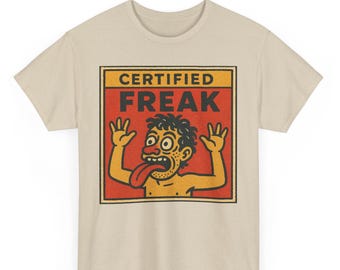 Pode incluir: Camiseta bege com um desenho de um personagem de desenho animado com cabelo selvagem, olhos esbugalhados e uma língua comprida. O personagem está dentro de uma caixa vermelha com as palavras "CERTIFIED FREAK".
