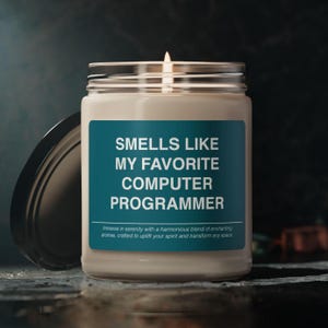 以下が含まれることがあります： ガラスの瓶に入った白いキャンドルで、ターコイズブルーのラベルには「Smells Like My Favorite Computer Programmer」と書かれています。ラベルには、「Immerse in serenity with a harmonious blend of enchanting aromas, crafted to uplift your spirit and transform any space.」というテキストも含まれています。