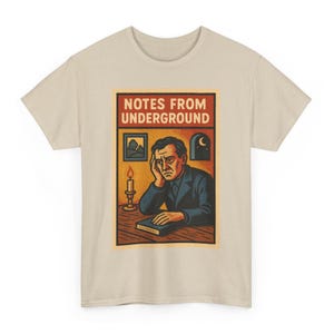 May include: Beige t-shirt featuring a rectangular graphic with the text "NOTES FROM UNDERGROUND." The graphic depicts a man resting his head on his hand, a candle, a window with a moon, and a landscape painting. The shirt is made of a soft material.