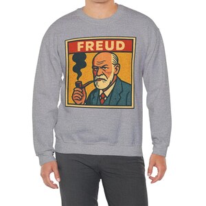 Może przedstawiać: Szara bluza z grafiką przedstawiającą Zygmunta Freuda palącego fajkę. Grafika znajduje się w prostokątnej ramce z napisem "FREUD" w czerwonych literach u g&oacute;ry. Obraz Freuda jest na ż&oacute;łtym tle.