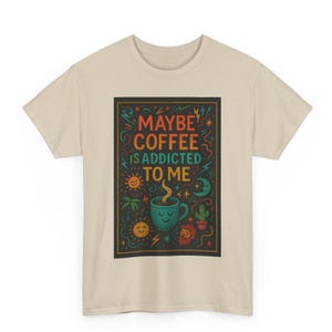 Puede incluir: Camiseta beige con un gráfico colorido que dice "MAYBE COFFEE IS ADDICTED TO ME". El diseño incluye una taza de café, un sol, una luna y otras ilustraciones. La camiseta está hecha de un material suave.