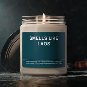 Peut inclure: Une bougie blanche dans un pot en verre avec une étiquette bleu sarcelle qui indique "SMELLS LIKE LAOS". L'étiquette comprend également le texte "Plongez dans la sérénité avec un mélange harmonieux d'arômes enchanteurs, conçus pour élever votre esprit et transformer n'importe quel espace."