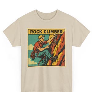 May include: Beige t-shirt featuring a vintage-style graphic of a rock climber. The illustration shows a climber on a colorful rock face with the words "ROCK CLIMBER" in a yellow box. The climber is wearing a red shirt, blue pants, and a yellow helmet.