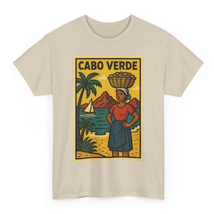 May include: Beige t-shirt featuring a rectangular graphic with the text "CABO VERDE." The graphic depicts a woman carrying a basket on her head, palm trees, a sailboat, and mountains. The woman wears a red shirt and blue skirt.