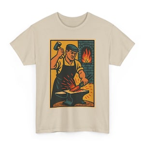 May include: Beige t-shirt featuring a rectangular graphic of a blacksmith at work. The illustration shows a blacksmith in a yellow shirt and black apron, hammering a piece of metal on an anvil. A brick forge with flames is in the background.