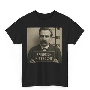 May include: Black t-shirt featuring a sepia-toned image of Friedrich Nietzsche holding a sign with his name. The image is a mugshot with height markings in the background. The shirt is a crew neck.