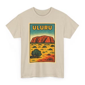 May include: Beige t-shirt with a rectangular graphic displaying the word "ULURU" in yellow. The graphic shows a red rock formation, green bushes, and a yellow and orange landscape, reminiscent of a vintage travel poster.