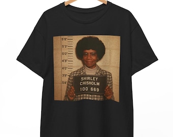 May include: Black t-shirt with a photo of Shirley Chisholm, a politician, in a mugshot style. The photo shows her in a plaid jacket and holding a sign that reads "Shirley Chisholm 100 669".