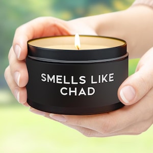 May include: A black candle with a white label that reads "Smells Like Chad". The candle is lit and the flame is visible.