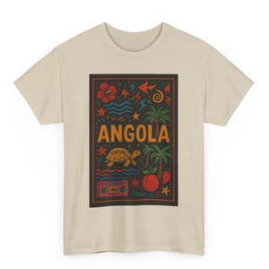 May include: Beige t-shirt with a rectangular graphic featuring the word "ANGOLA" in orange. The graphic includes a turtle, palm trees, a boombox, and other colorful illustrations. The shirt is made of a soft, comfortable material.