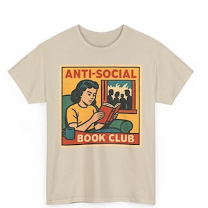 May include: Beige t-shirt featuring a retro-style graphic. The graphic depicts a person reading a book with the words "ANTI-SOCIAL BOOK CLUB" in a rectangular frame. The design includes a person, a window, and a coffee cup.