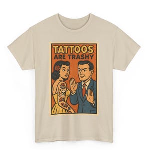 May include: Beige t-shirt featuring a retro-style graphic. The graphic shows a woman with tattoos and a man gesturing with his hands, with the text "TATTOOS ARE TRASHY" in a bold font. The design has a vintage comic book aesthetic.