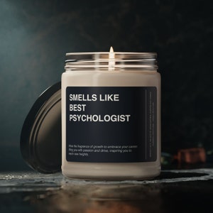 May include: A lit, white scented candle in a glass jar with a black label that reads "SMELLS LIKE BEST PSYCHOLOGIST". The candle's lid is off to the side. The text below the title reads "Allow the fragrance of growth to embrace your career, filling you with passion and drive, inspiring you to reach new heights."