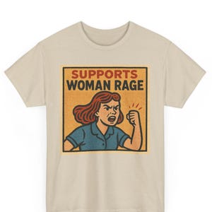 Puede incluir: Camiseta beige con un gráfico retro. El diseño presenta a una mujer con cabello rojo y un puño cerrado, con las palabras "SUPPORTS WOMAN RAGE" en negrita. El gráfico está sobre un fondo naranja texturizado.