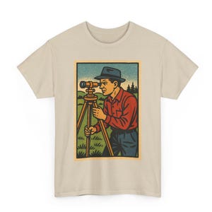 Pode incluir: Camiseta bege com um gráfico retangular que retrata um homem usando um instrumento de topografia. O homem usa uma camisa vermelha, calças azuis e um chapéu escuro. O fundo mostra um campo verde, céu azul e árvores.