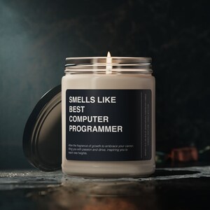 May include: A glass candle jar with a black label that reads "Smells Like Best Computer Programmer". The candle is lit and the jar is sitting on a dark surface.
