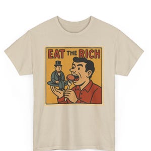 May include: Beige t-shirt with a retro-style graphic. The graphic features a man eating a small figure on a plate, with the text "EAT THE RICH" in a bold font. The design is outlined in black and set against a yellow background.
