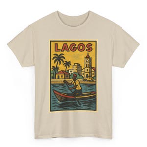 May include: Beige t-shirt featuring a rectangular graphic with the word "LAGOS" in red. The graphic depicts a person rowing a boat on blue water, with palm trees, buildings, and a church in the background. The shirt is a crew neck.
