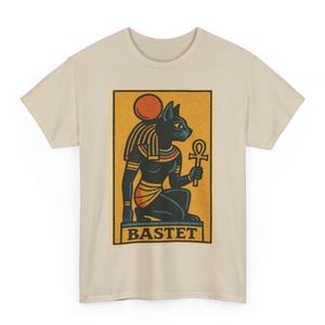 May include: Beige t-shirt with a rectangular graphic of the Egyptian goddess Bastet. The graphic shows a black cat-headed figure holding an ankh, with the word "BASTET" at the bottom. The design includes orange, yellow, and black elements.