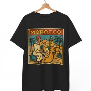 May include: Black t-shirt featuring a retro-style graphic of a person riding a camel through a desert landscape. The graphic includes the word "MOROCCO" in a rectangular frame, palm trees, and buildings.