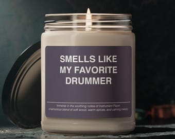 May include: A white candle in a glass jar with a purple label that reads "Smells Like My Favorite Drummer". The label also includes the text "Immerse in the soothing notes of Instrument Player. A harmonious blend of soft wood, warm spices, and calming melody."