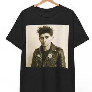 Op de afbeelding: Zwart T-shirt met een vintage foto van een man met stekelig haar, die een zwart leren jack draagt met studded details en twee badges. Een badge zegt "A.D. 1000" en de andere "A.H."