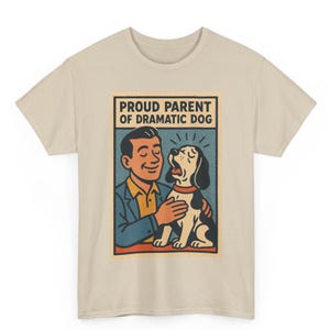 May include: Beige t-shirt featuring a retro-style graphic. The graphic depicts a man with a dog, with the text "PROUD PARENT OF DRAMATIC DOG" in a rectangular frame. The illustration has a vintage aesthetic with a limited color palette.