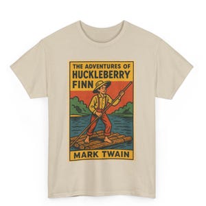 Puede incluir: Camiseta beige con un gráfico colorido de "Las aventuras de Huckleberry Finn". La ilustración muestra a un niño en una balsa, sosteniendo una pértiga, con el texto "The Adventures of Huckleberry Finn" y "Mark Twain".