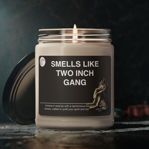 Pode incluir: Uma vela branca em um pote de vidro com um rótulo preto que diz "Smells Like Two Inch Gang". O rótulo também inclui um pequeno ícone de lua e um desenho de uma criatura semelhante a um lobo.