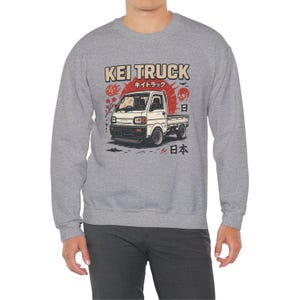 Pu&ograve; includere: Felpa grigia con grafica di un camion Kei bianco e testo giapponese. Il design include un sole rosso, un teschio e caratteri giapponesi. La felpa &egrave; in stile casual.