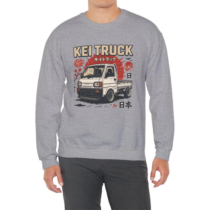Pu&ograve; includere: Felpa grigia con grafica di un camion Kei bianco e testo giapponese. Il design include un sole rosso, un teschio e caratteri giapponesi. La felpa &egrave; in stile casual.