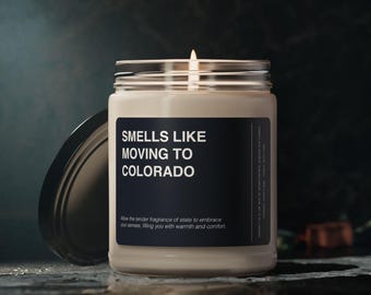 Op de afbeelding: Een kaars in een glazen pot met een zwart label waarop "Smells Like Moving to Colorado" staat. Het label bevat ook de tekst "Allow the tender fragrance of state to embrace your senses, filling you with warmth and comfort."