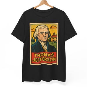 Puede incluir: Camiseta negra con un gráfico vintage de Thomas Jefferson. El diseño incluye un retrato de Jefferson, un edificio y el texto "THOMAS JEFFERSON" en una pancarta roja. El fondo tiene tonos amarillos y naranjas.