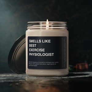 Può includere: Una candela accesa in un barattolo di vetro con un'etichetta nera che recita "SMELLS LIKE BEST EXERCISE PHYSIOLOGIST". La candela ha un coperchio argentato e si trova su una superficie scura.