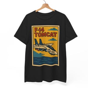 May include: Black t-shirt featuring a vintage-style graphic of an F-14 Tomcat fighter jet. The design includes the text "F-14 TOMCAT" in a retro font, with the jet flying over water. The graphic is in shades of blue, yellow, and orange.