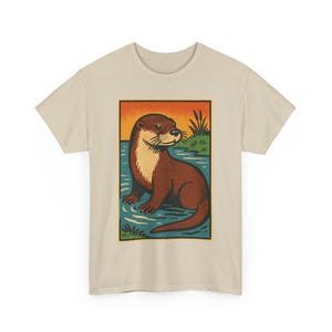 May include: Beige t-shirt with a rectangular graphic depicting an otter in a river. The otter is brown with a cream chest, set against an orange and blue backdrop. The design has a vintage, illustrated style.