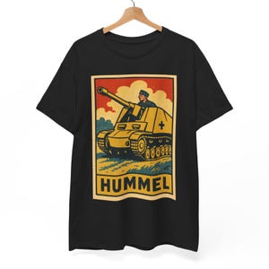 以下が含まれることがあります： 黒いTシャツ。レトロなデザインで、黄色い戦車と兵士が赤と黄色の背景に描かれています。「HUMMEL」の文字が黄色で下部にプリントされています。クラシックなミリタリーテーマのグラフィックTシャツ。