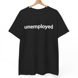 May include: Black t-shirt with the word "unemployed" printed in white, displayed on a wooden hanger. The shirt has short sleeves and a crew neck.