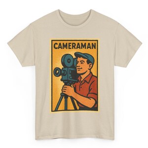 Pode incluir: Camiseta bege com um gráfico retrô de um cinegrafista segurando uma câmera de filme vintage em um tripé. O gráfico está em uma moldura retangular com a palavra "CAMERAMAN" na parte superior. O cinegrafista está vestindo uma camisa vermelha e um boné azul.