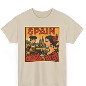 以下が含まれることがあります： ベージュ色のTシャツ。黄色と赤で「SPAIN」の文字が入ったグラフィック。グラフィックは闘牛士、赤いバラを持つ女性、雄牛、教会を描いています。デザインはヴィンテージスタイルです。